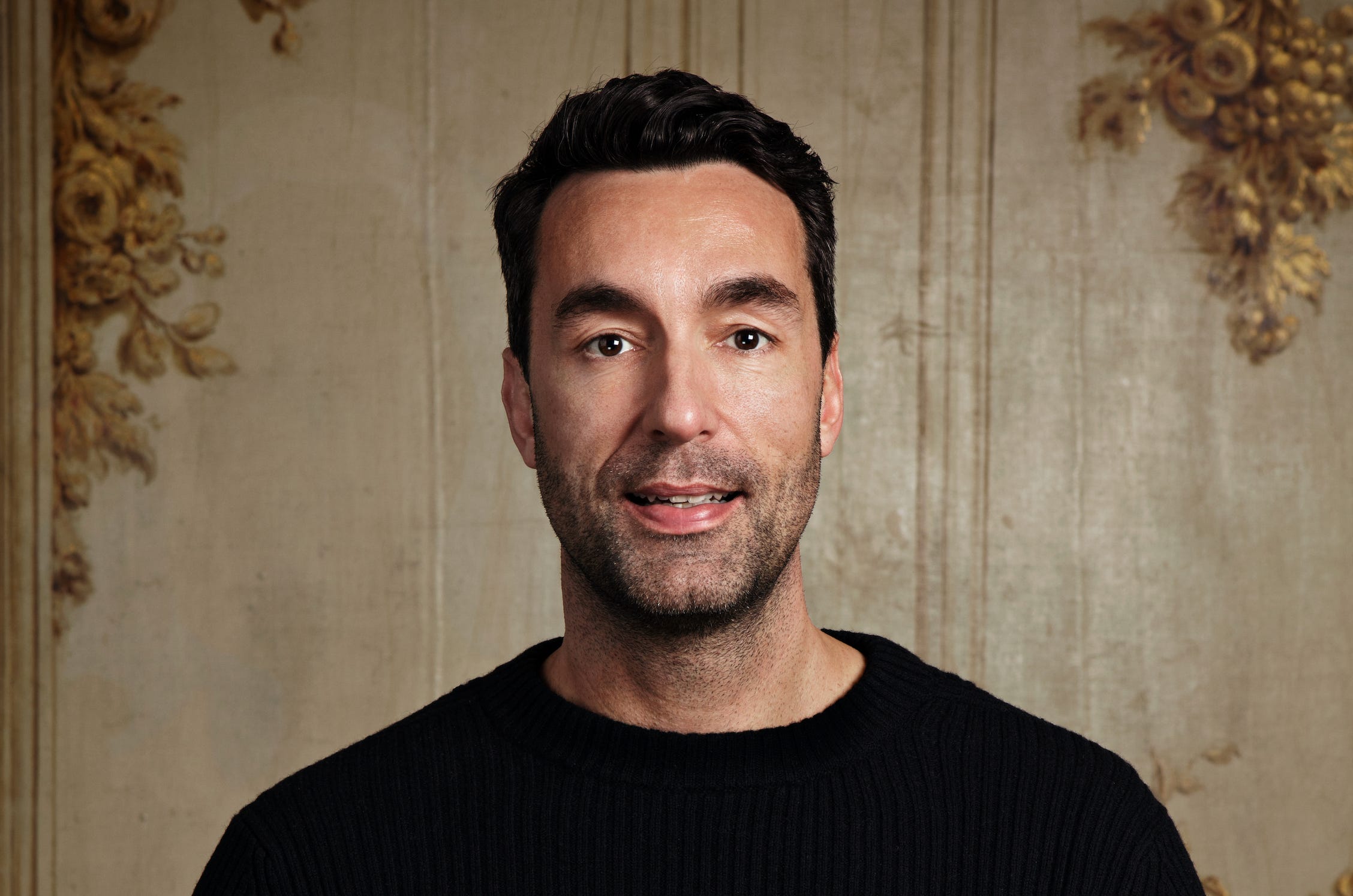 Une personne avec des cheveux noirs et un pull noir devant un mur vintage décoré.