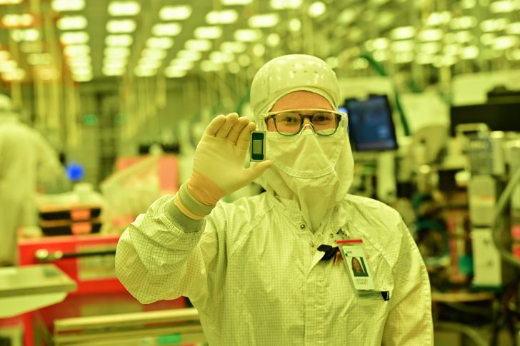 A person in a cleanroom suit holds a small, unbranded microchip in a manufacturing facility.