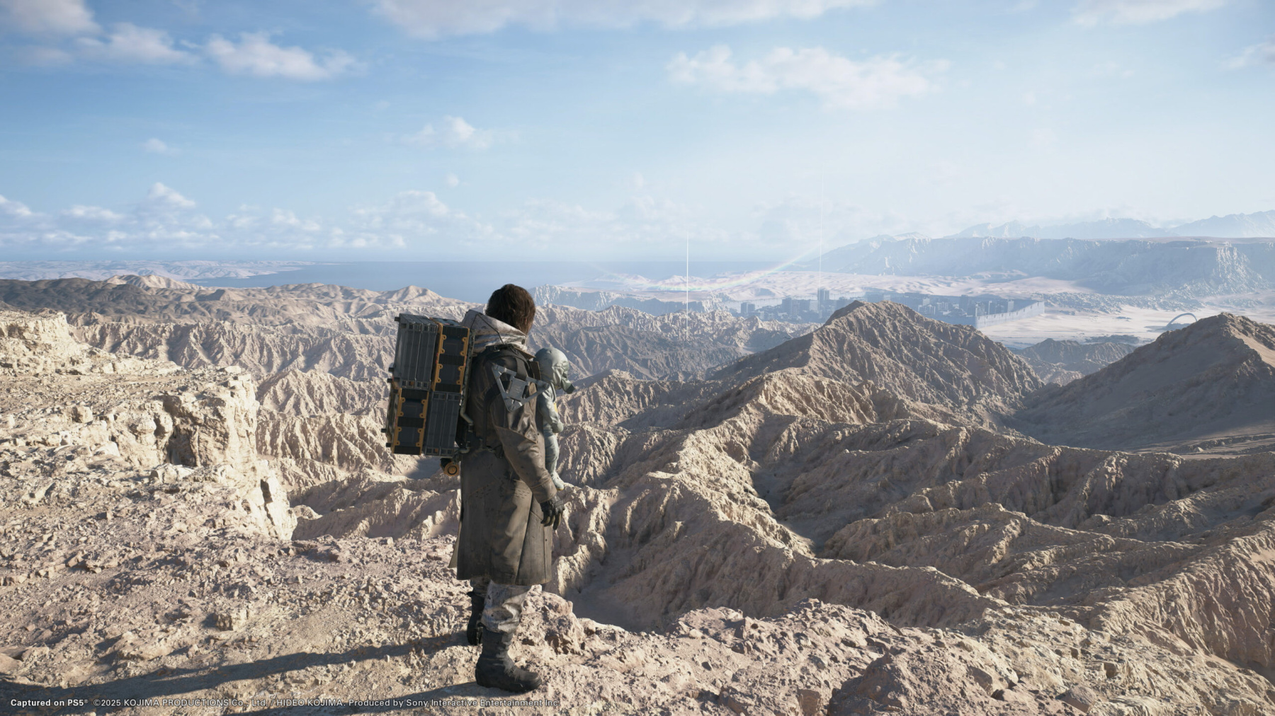 Un personnage de 'Death Stranding' sur un montagne rocheuse, avec une vue sur un paysage accidenté.