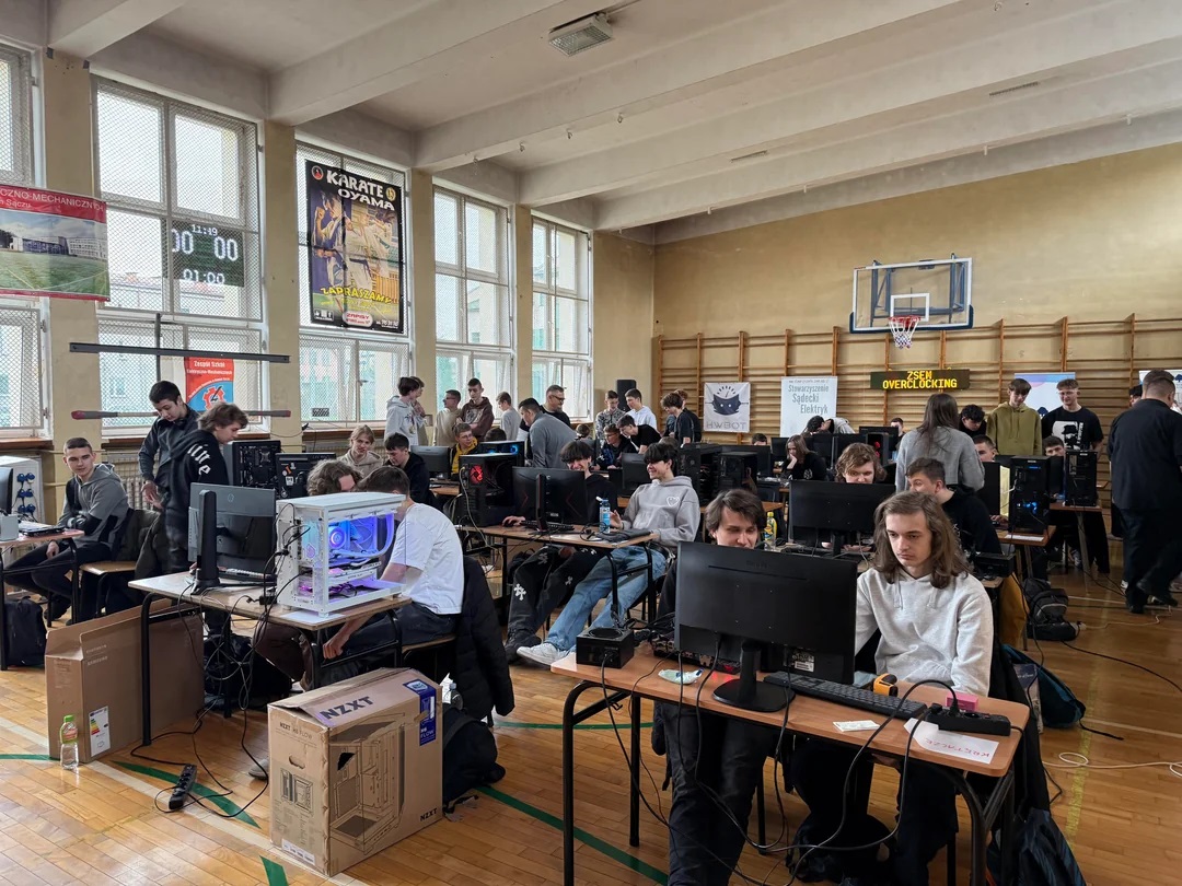 Participants in a school gym competing in a 'ZSEM Overclocking' event, with gaming PCs, monitors, and an NZXT case box visible.