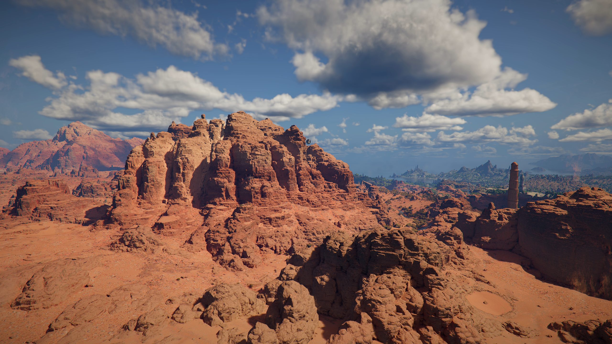 A detailed landscape of a desert with red rock formations, a tall tower-like structure, and a distant mountain range under a partly cloudy sky, from an open-world video game.
