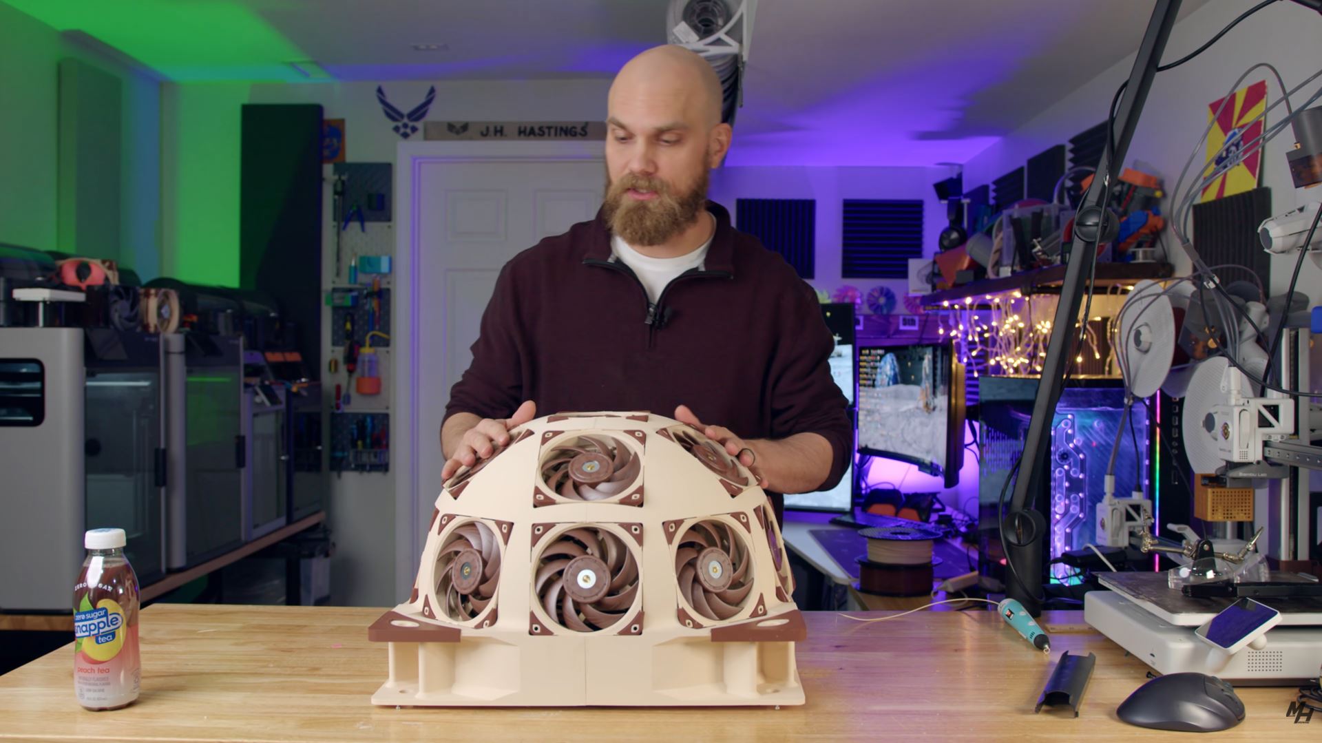 A person stands in a workshop behind a dome-shaped structure with multiple Noctua cooling fans, next to a bottle of Snapple peach tea on a table.