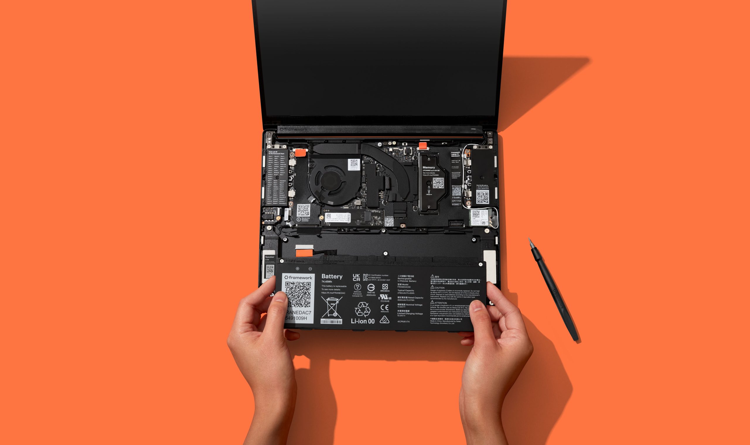 A person's hands are holding a Framework laptop battery labeled 'Li-ion 00,' showing the internal components of the device on an orange background.