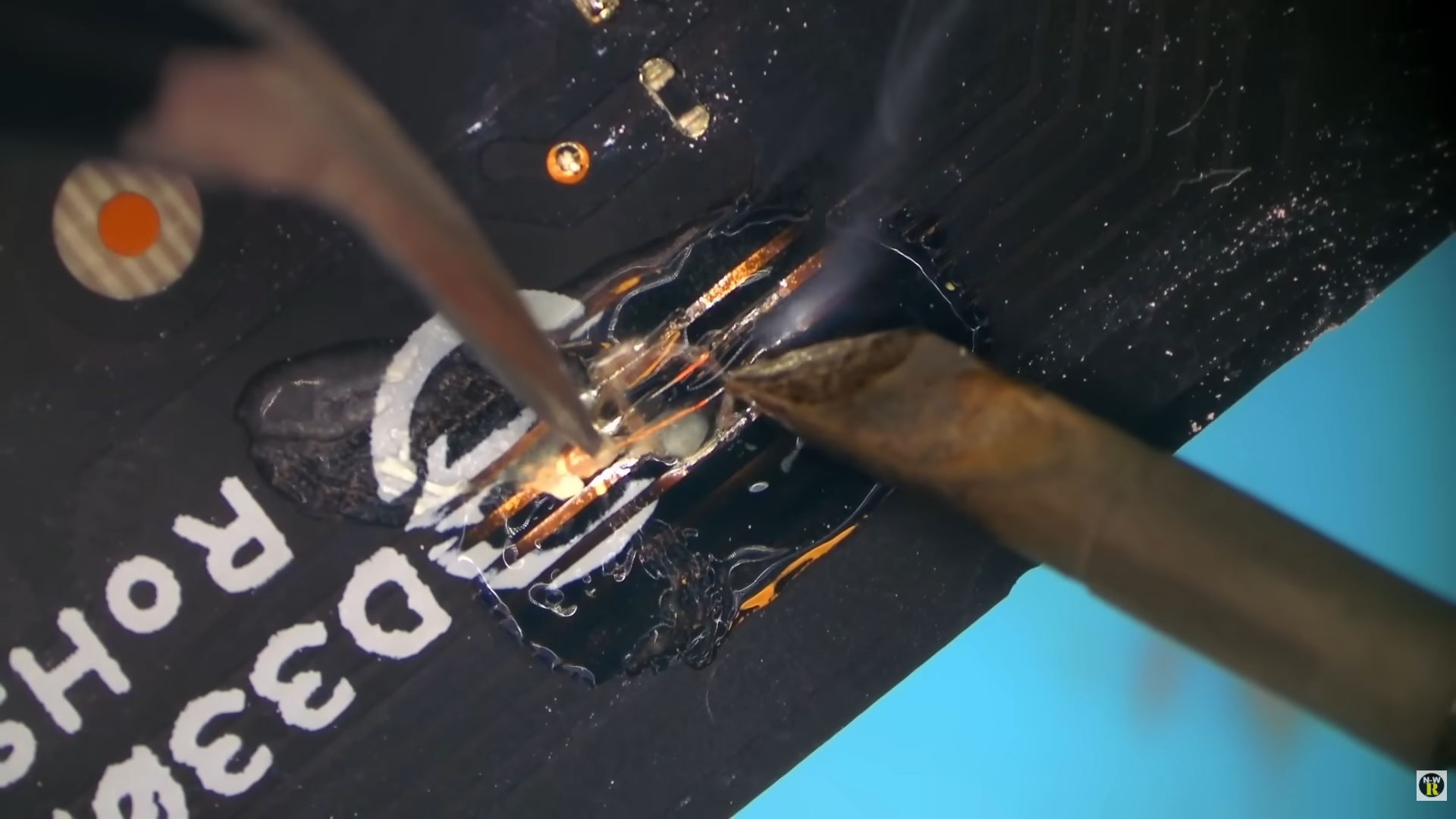 A soldering iron and tweezers are being used to repair a damaged circuit board labeled with 'ROHS' and '003ED'.
