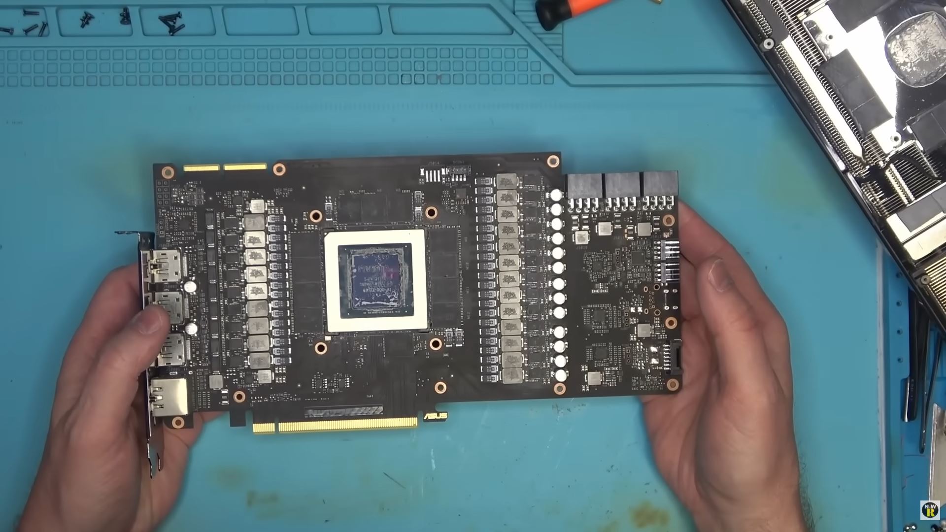 A person holds an ASUS graphics card with an exposed GPU and various visible components on a blue work mat.
