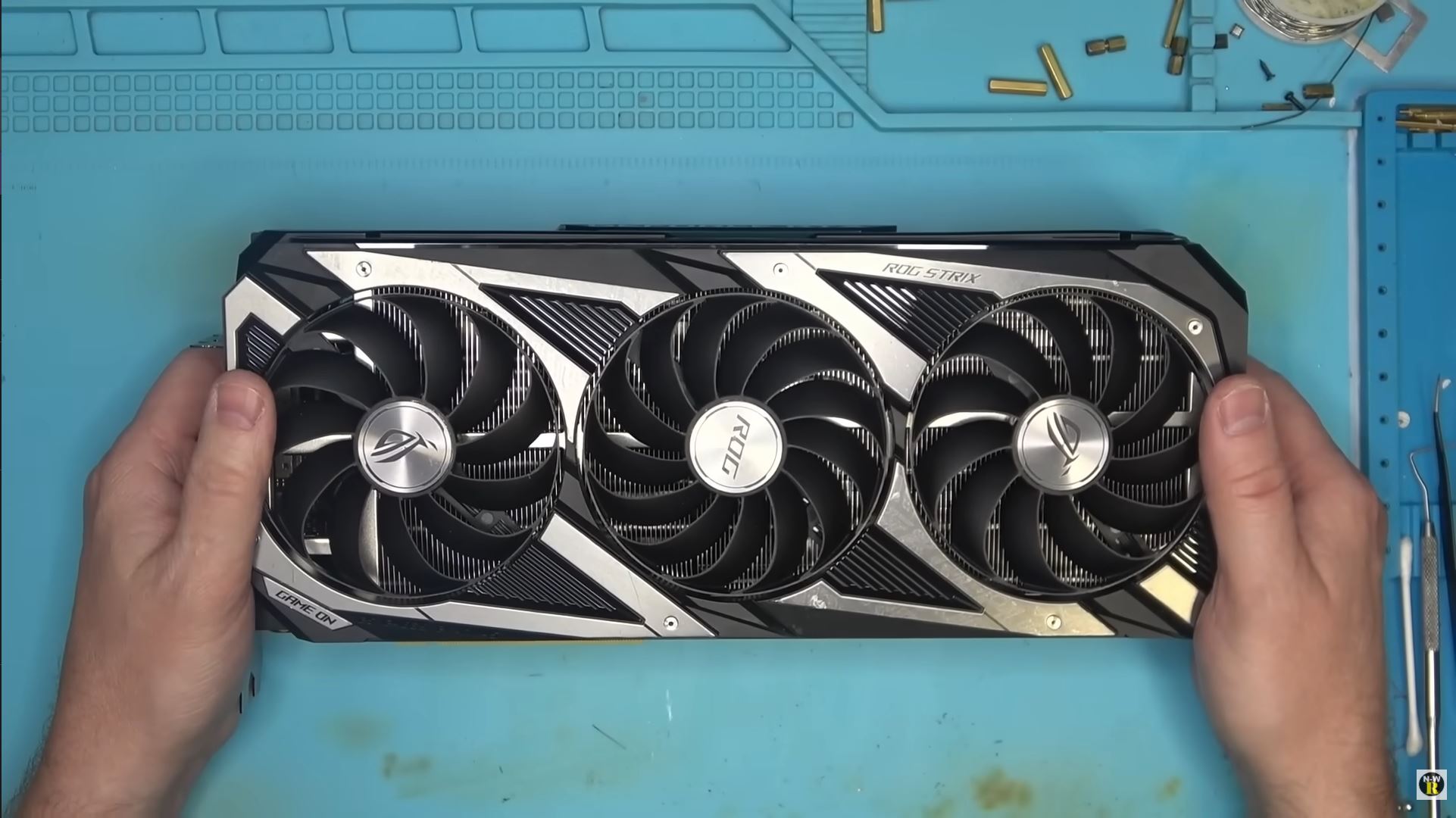 An ASUS ROG STRIX graphics card with three cooling fans is held by hands over a blue work mat.