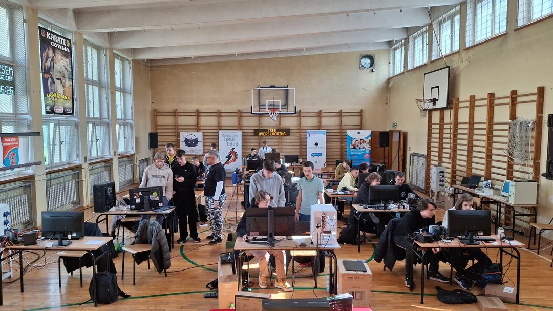 A gymnasium filled with people working on computers under a 'ZSEH OVERCLOCKING' banner, surrounded by branded posters and tables with various electronic equipment.