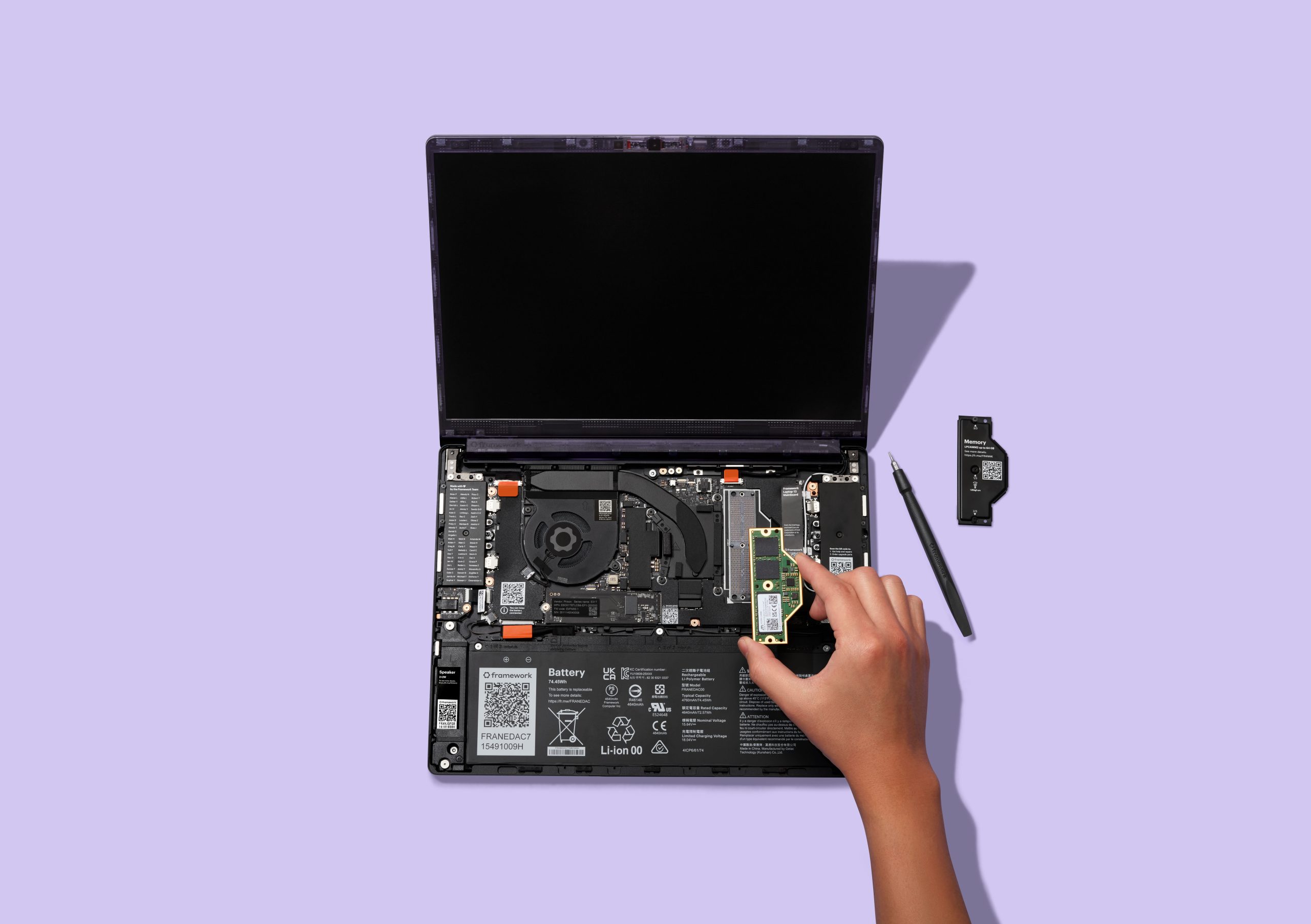 A hand installs a Framework-branded memory module into an open Framework laptop with a visible internal battery labeled 'FRANEDC47'.