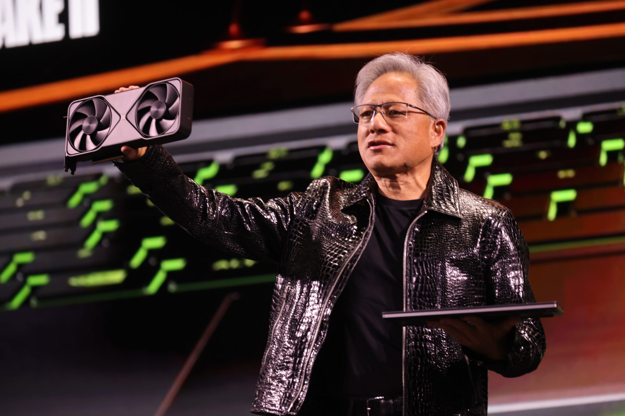 A person in a shiny jacket holds up a graphics card on stage with a presentation backdrop.