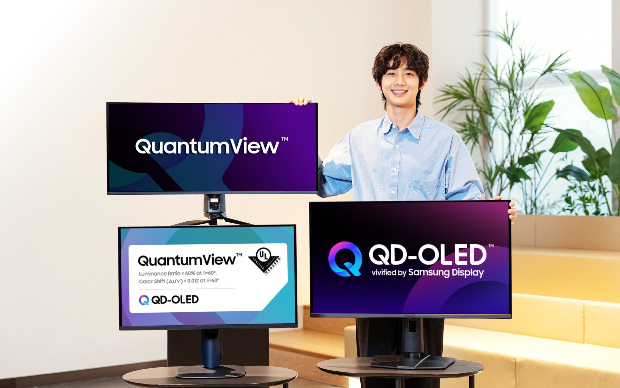 A person stands next to Samsung QuantumView monitors with the text 'QuantumView' and 'QD-OLED vivified by Samsung Display' visible on the screens.
