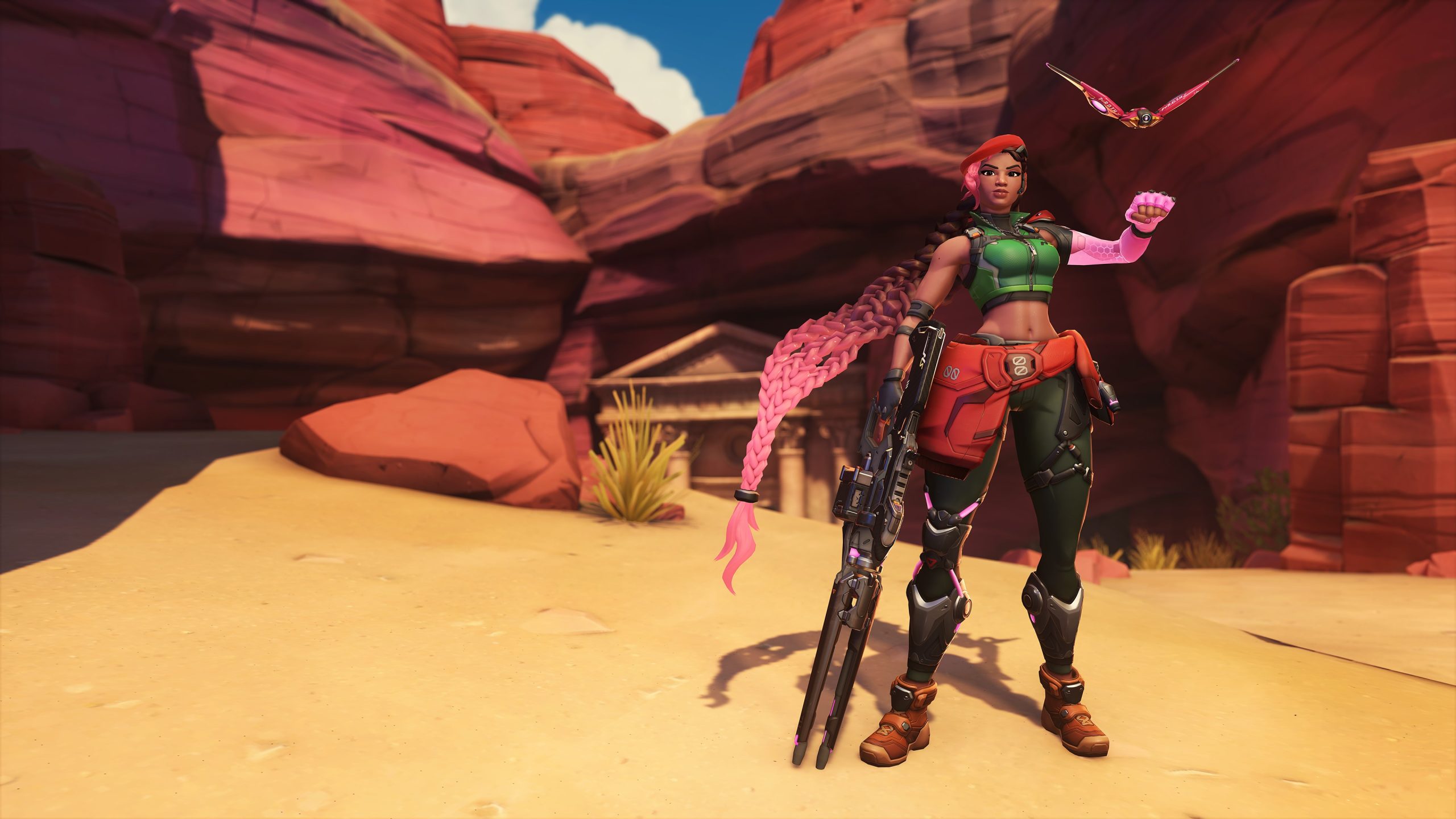 A character in 'Overwatch' stands in a desert canyon holding a weapon with a robotic bird hovering above.
