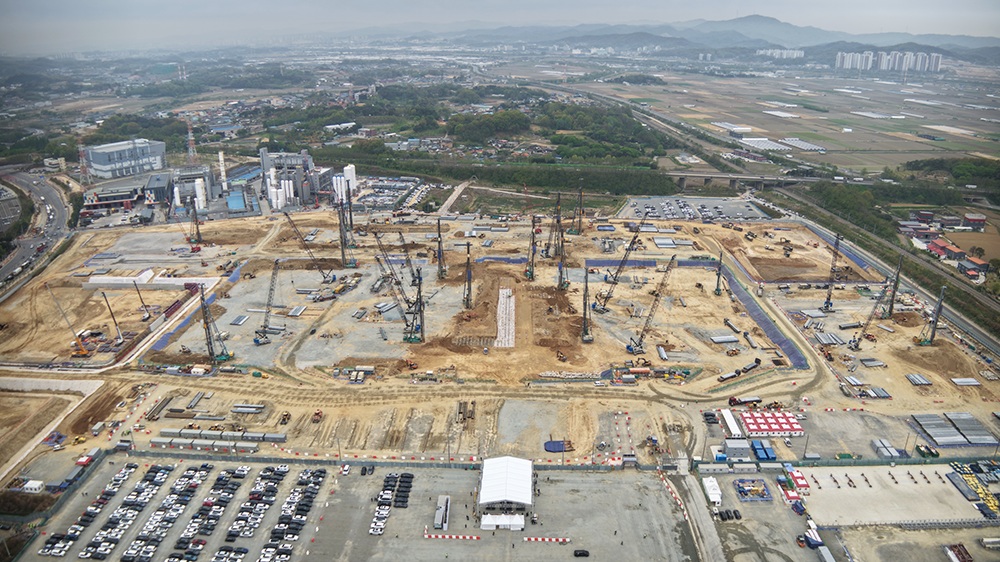 An aerial view of a large construction site with multiple cranes and a parking lot with numerous vehicles, surrounded by rural and industrial areas.