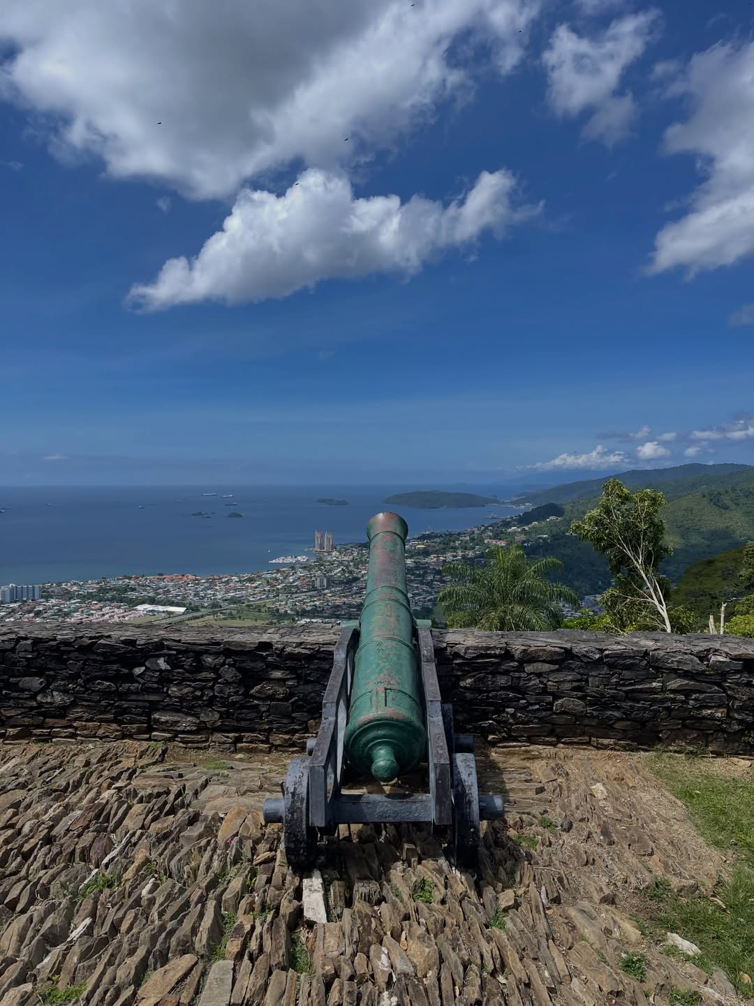 An old cannon is mounted on a stone platform overlooking a scenic coastal cityscape under a blue sky with clouds.