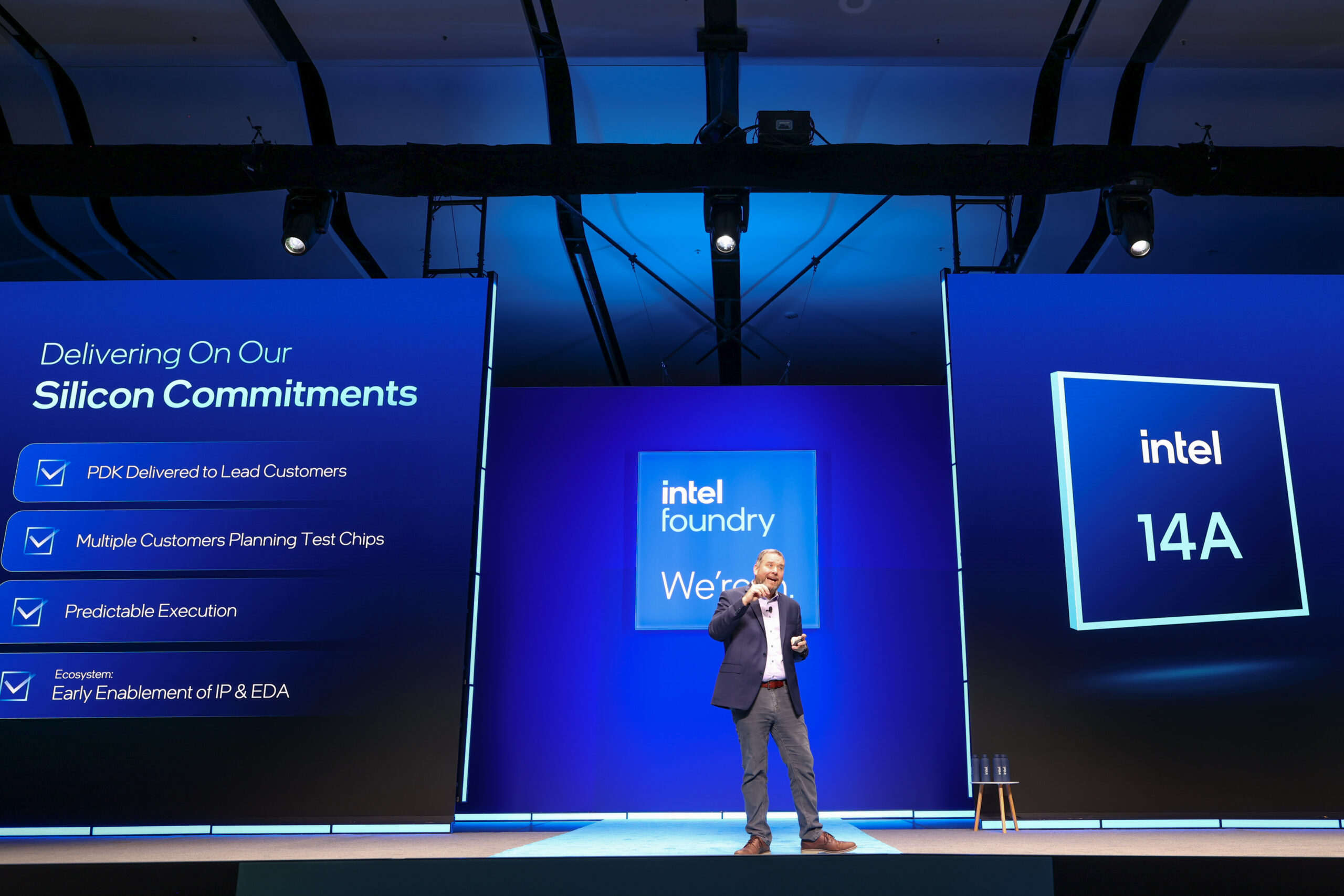 A presenter speaks on stage in front of screens displaying 'Delivering On Our Silicon Commitments' and 'intel 14A' at an Intel Foundry event.