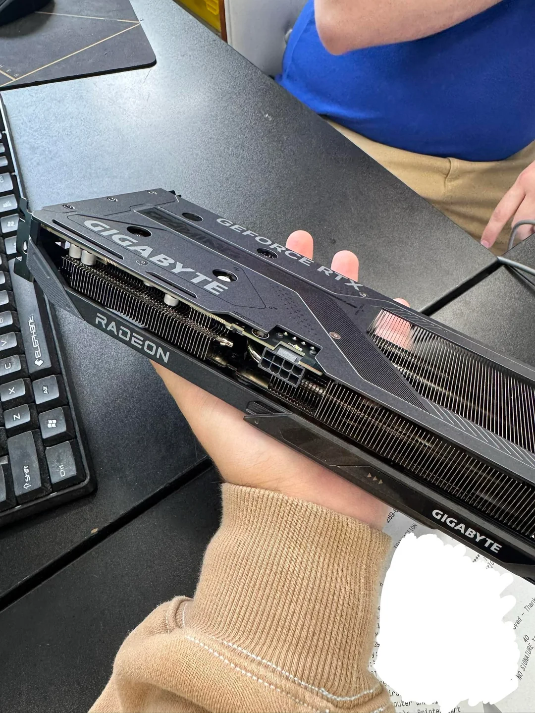 A person holding a Gigabyte graphics card with a visible 'GeForce RTX' and 'Radeon' branding on a desk next to a keyboard.