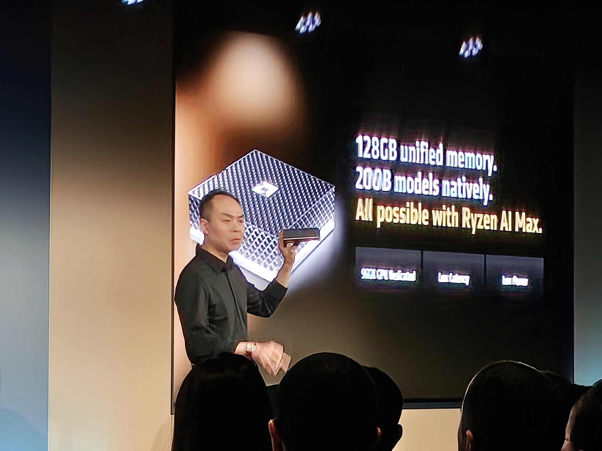 A presenter on stage holding a chip next to a slide displaying the text '128GB unified memory, 200B models natively, All possible with Ryzen AI Max.'