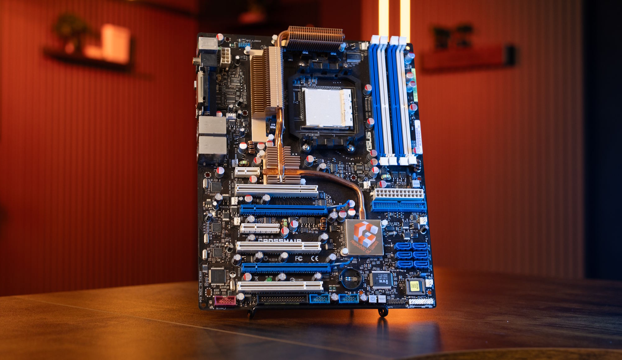 An ASUS motherboard with multiple PCI slots, blue RAM slots, and copper heat sinks, displayed upright on a wooden surface.