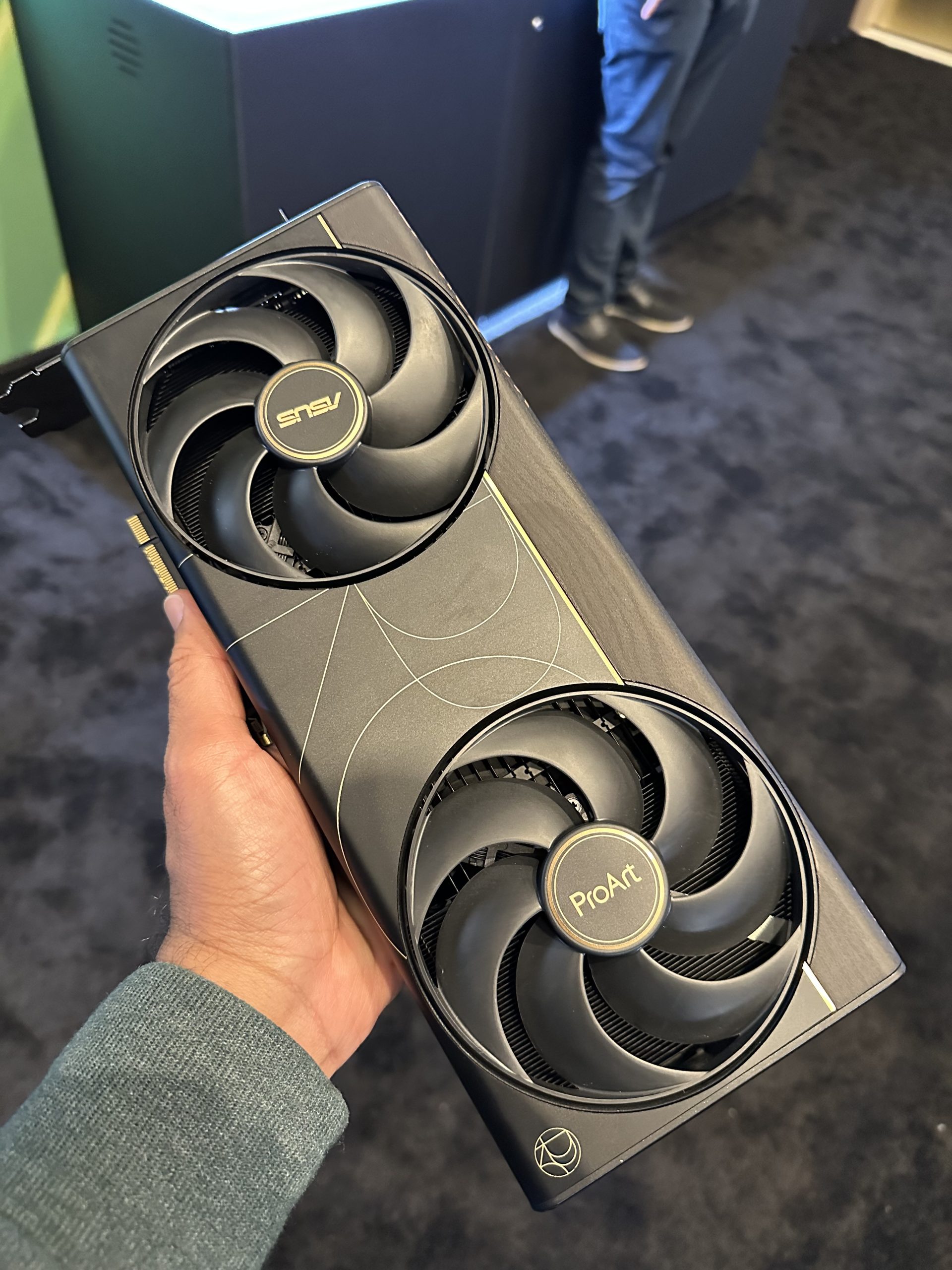 A hand holding an ASUS ProArt graphics card with dual fans, visible branding, and a geometric design pattern.