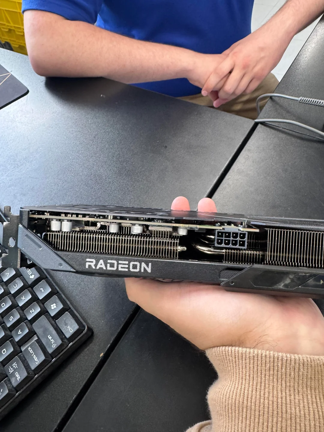 A person holding a Radeon graphics card with visible cooling components and an 8-pin power connector, beside a keyboard.
