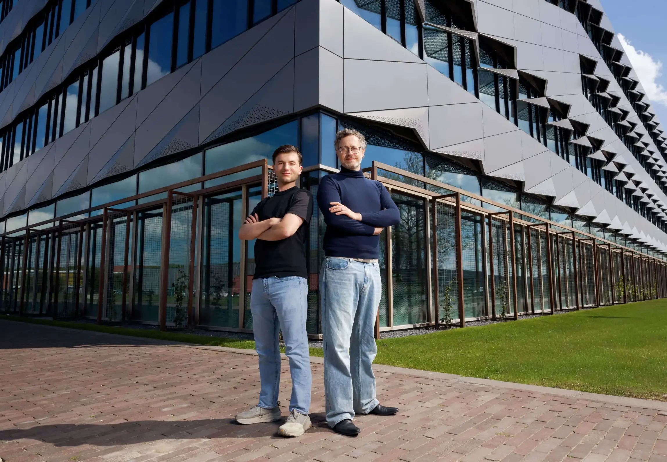Two individuals stand with arms crossed in front of a modern office building with geometric architectural features and large glass windows.