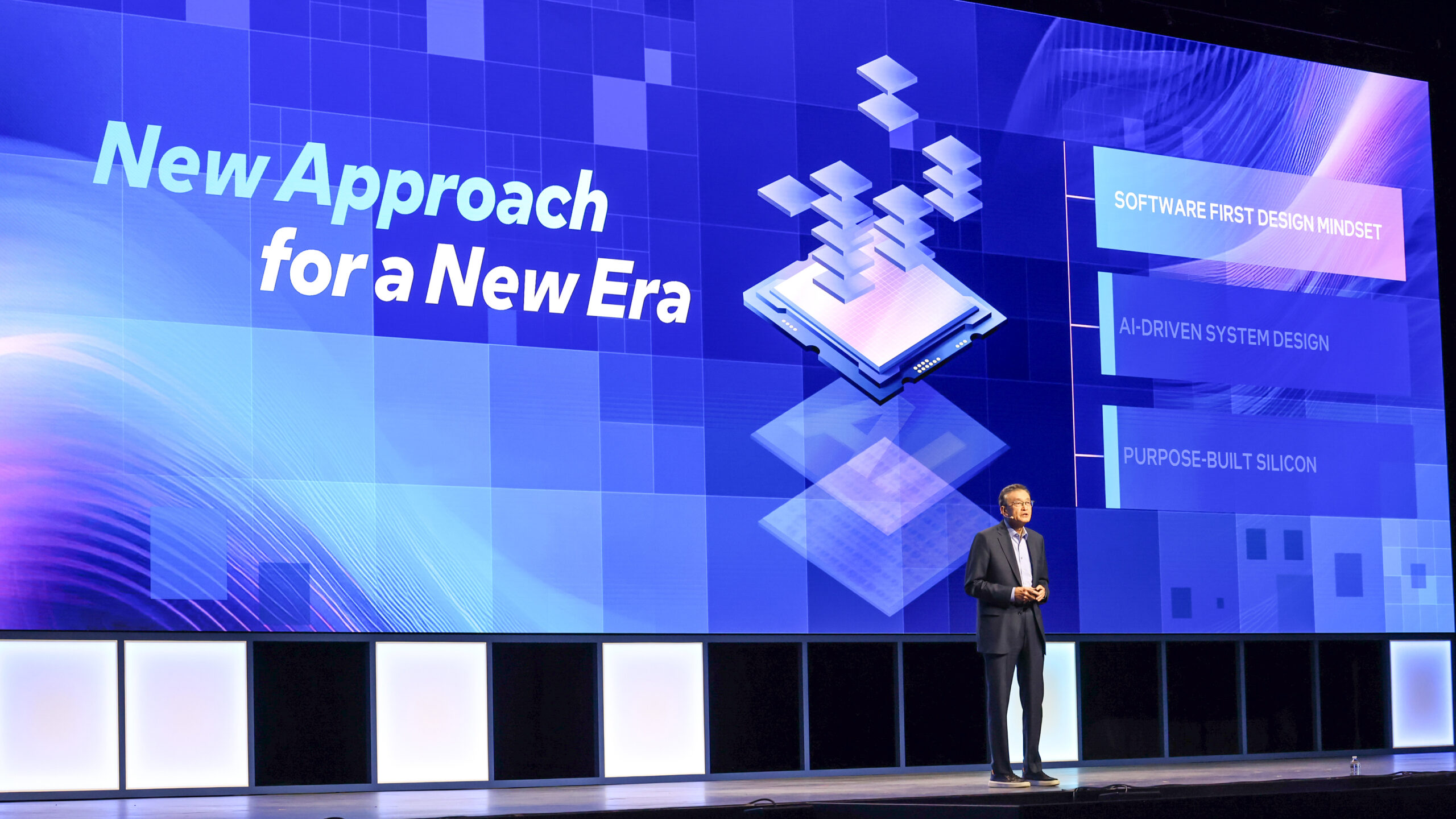 A person stands on stage in front of a screen displaying New Approach for a New Era with project titles like AI-DRIVEN SYSTEM DESIGN.