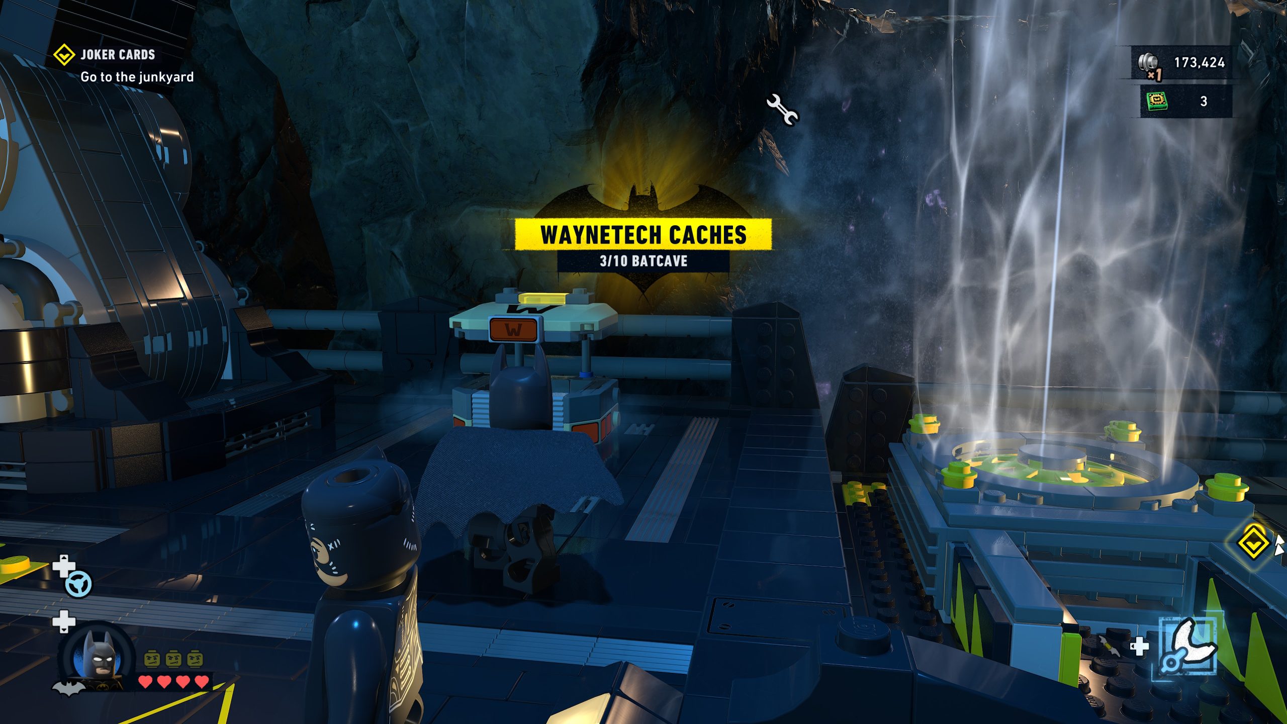 Dans le jeu 'LEGO Batman Legacy of the Dark Knight,' Batman dans la Batcave, texte 'WAYNETECH CACHES 3/10 BATCAVE' et 'JOKER CARDS Go to the junkyard.'