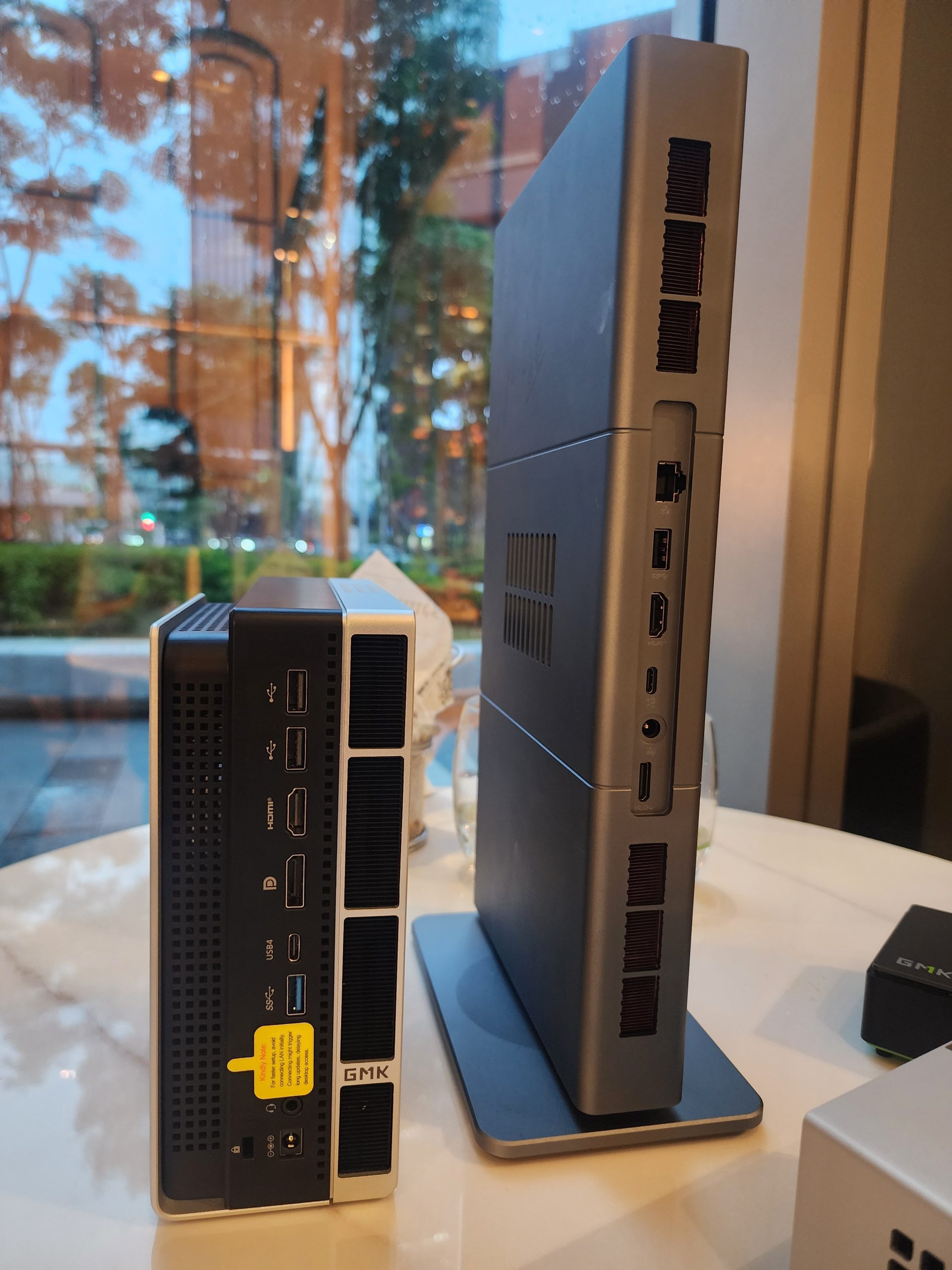 Un mini PC GMK NucBox 7 placé à côté d'un PC de bureau fin et non marqué, illustrant leurs designs compacts et leurs différents ports.