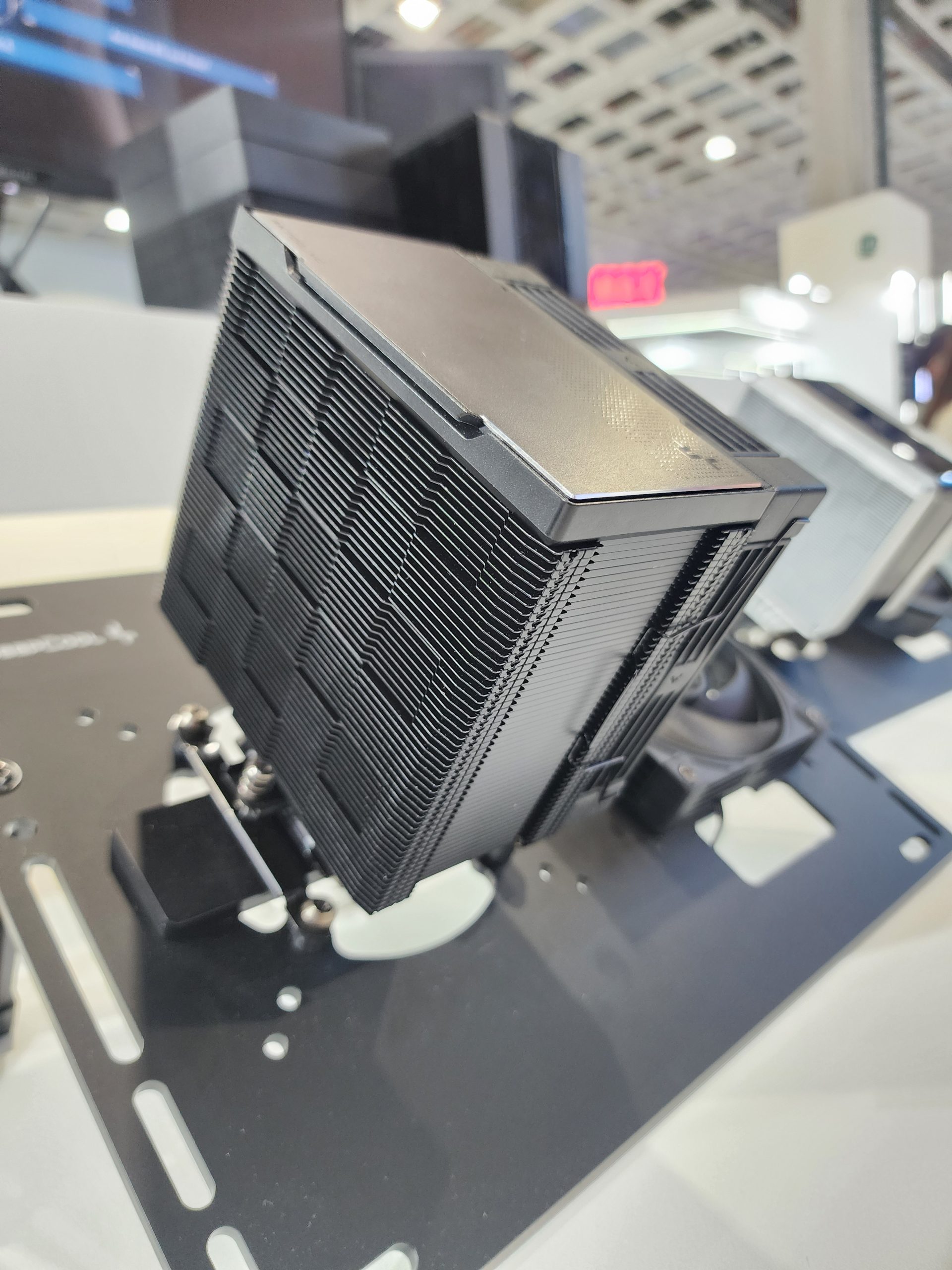 Un gros refroidisseur CPU noir avec des ailettes empilées est exposé sur un support de montage dans un stand d'exposition.