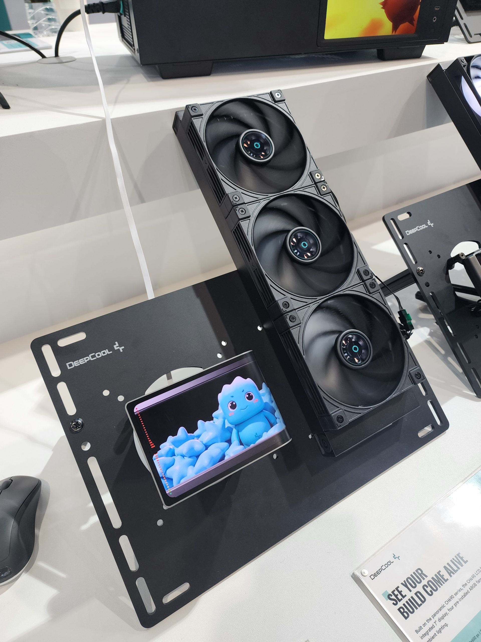 Un système de refroidissement DeepCool présenté avec une configuration à trois ventilateurs à côté d'un personnage animé bleu sur un écran avec le texte 'DeepCool' visible.