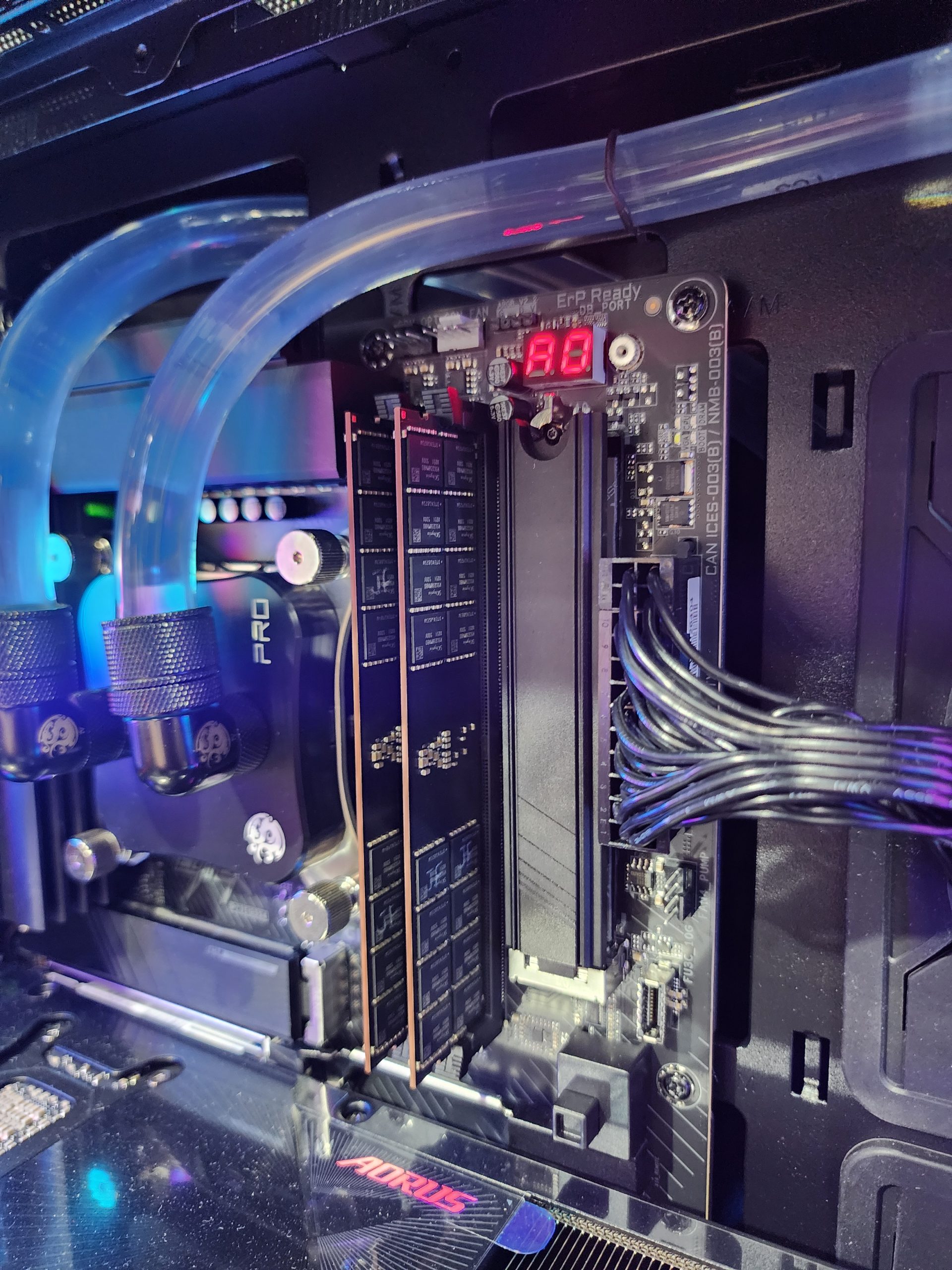 Un PC personnalisé avec une carte mère AORUS, des tubes éclairés visibles, un écran affichant '98' et des modules RAM installés.