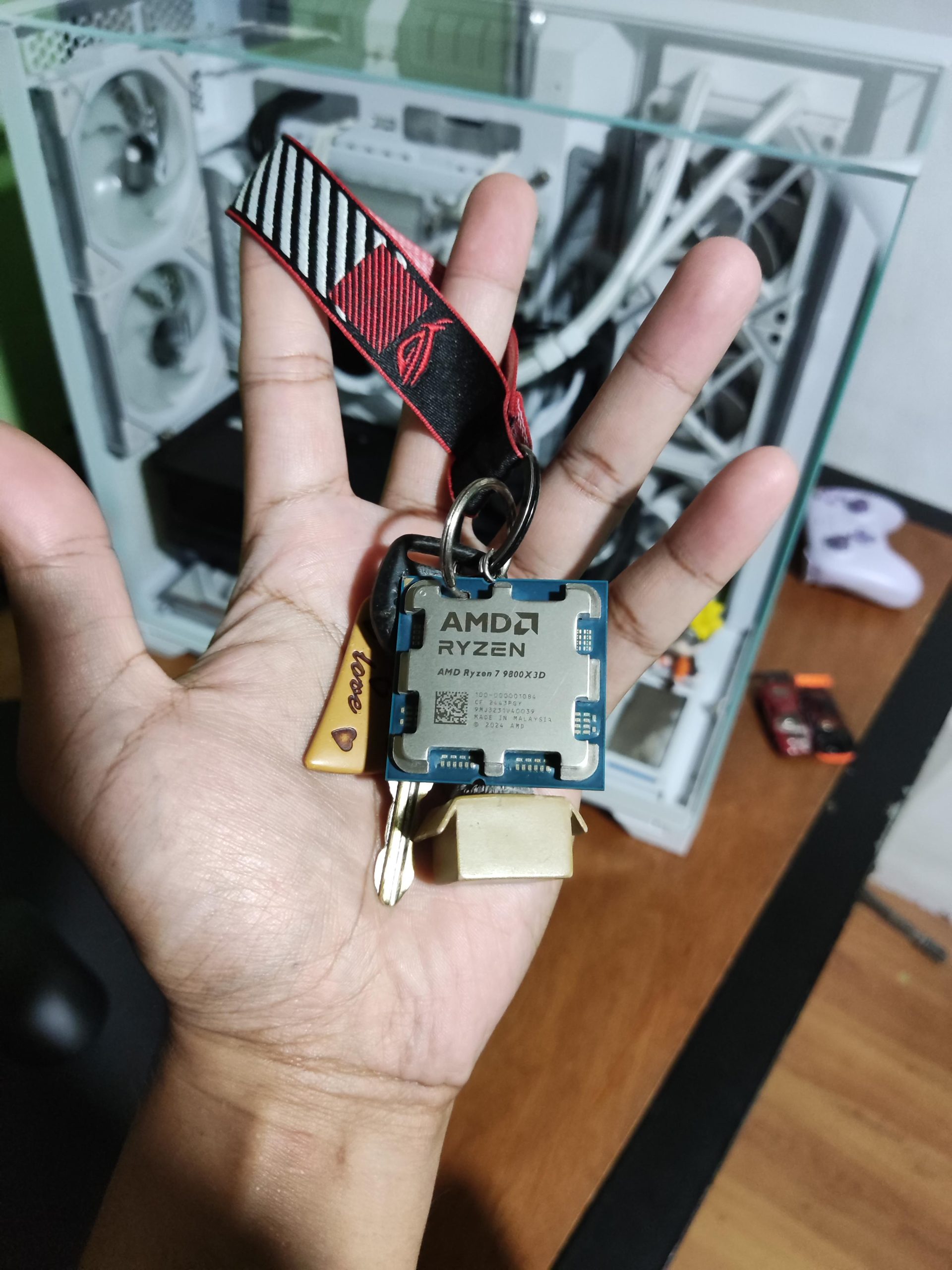 Une personne tenant un processeur AMD Ryzen 7 7800X3D monté en porte-clés, avec un PC gaming en arrière-plan.
