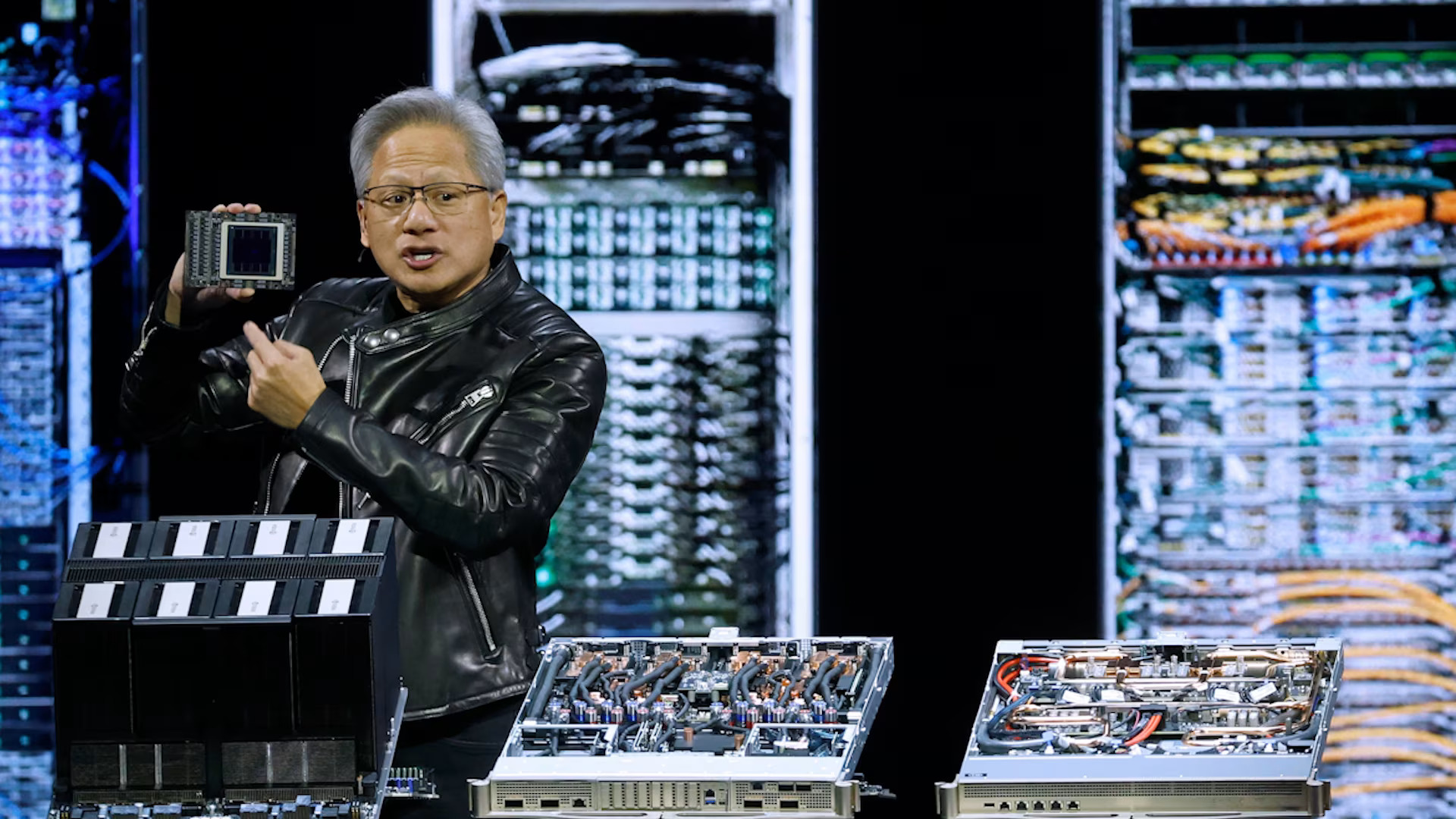 Homme présentant un GPU NVIDIA sur scène avec des baies de serveurs en arrière-plan.