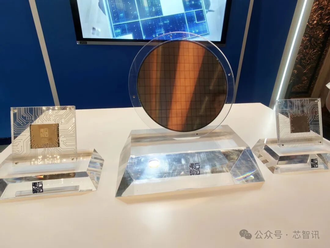 Un écran présentant une galette de silicium et deux puces dans des blocs transparents, avec des motifs de circuits visibles sur une table.