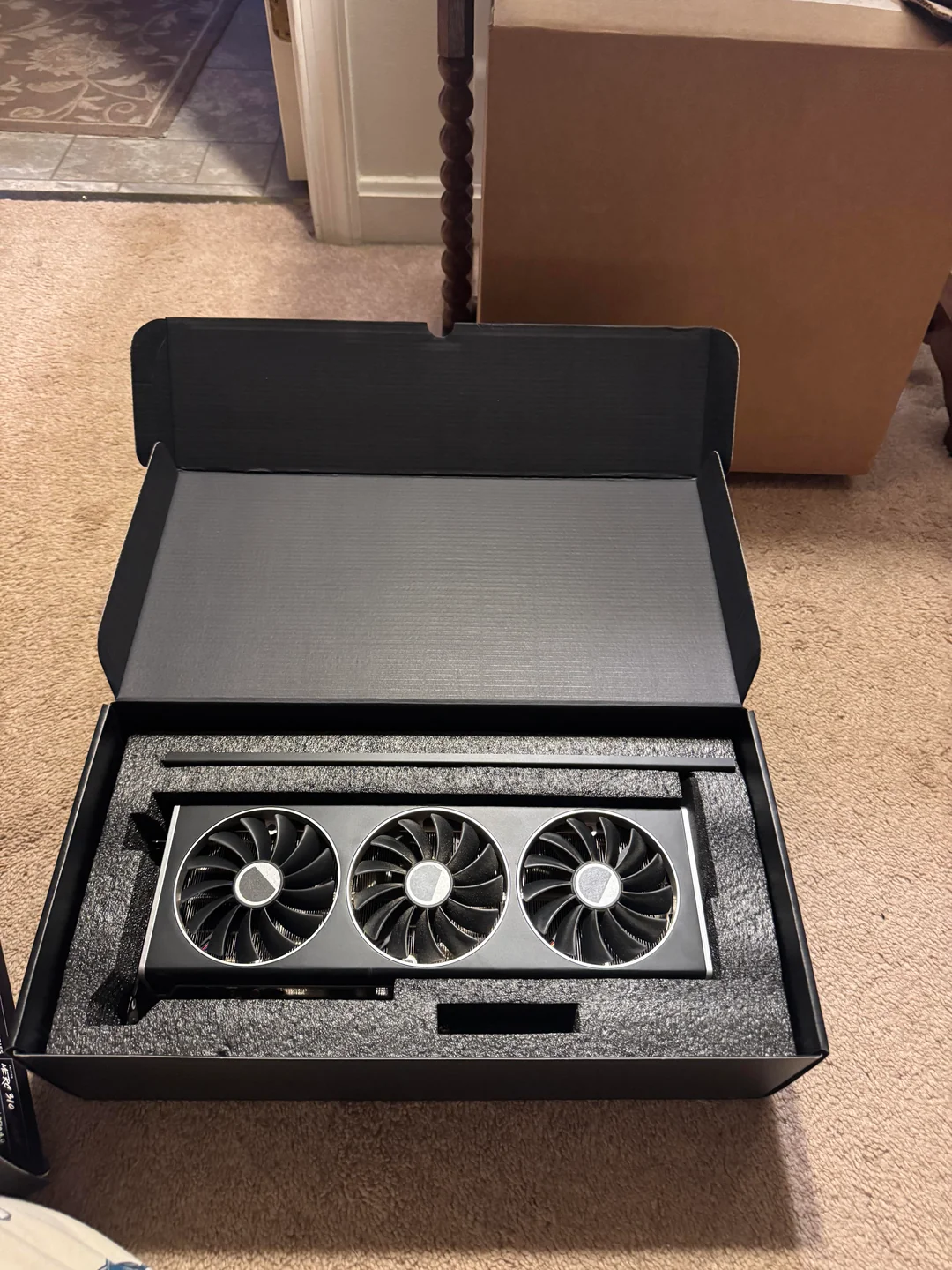 Photographie de la carte graphique XFX Radeon RX 7900 XT à trois ventilateurs, dans sa boîte ouverte, posée sur un sol.
