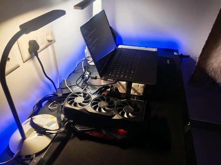 Un setup avec un portable non marqué et une carte graphique AMD Radeon RX à trois ventilateurs sur un bureau, sous une lampe.