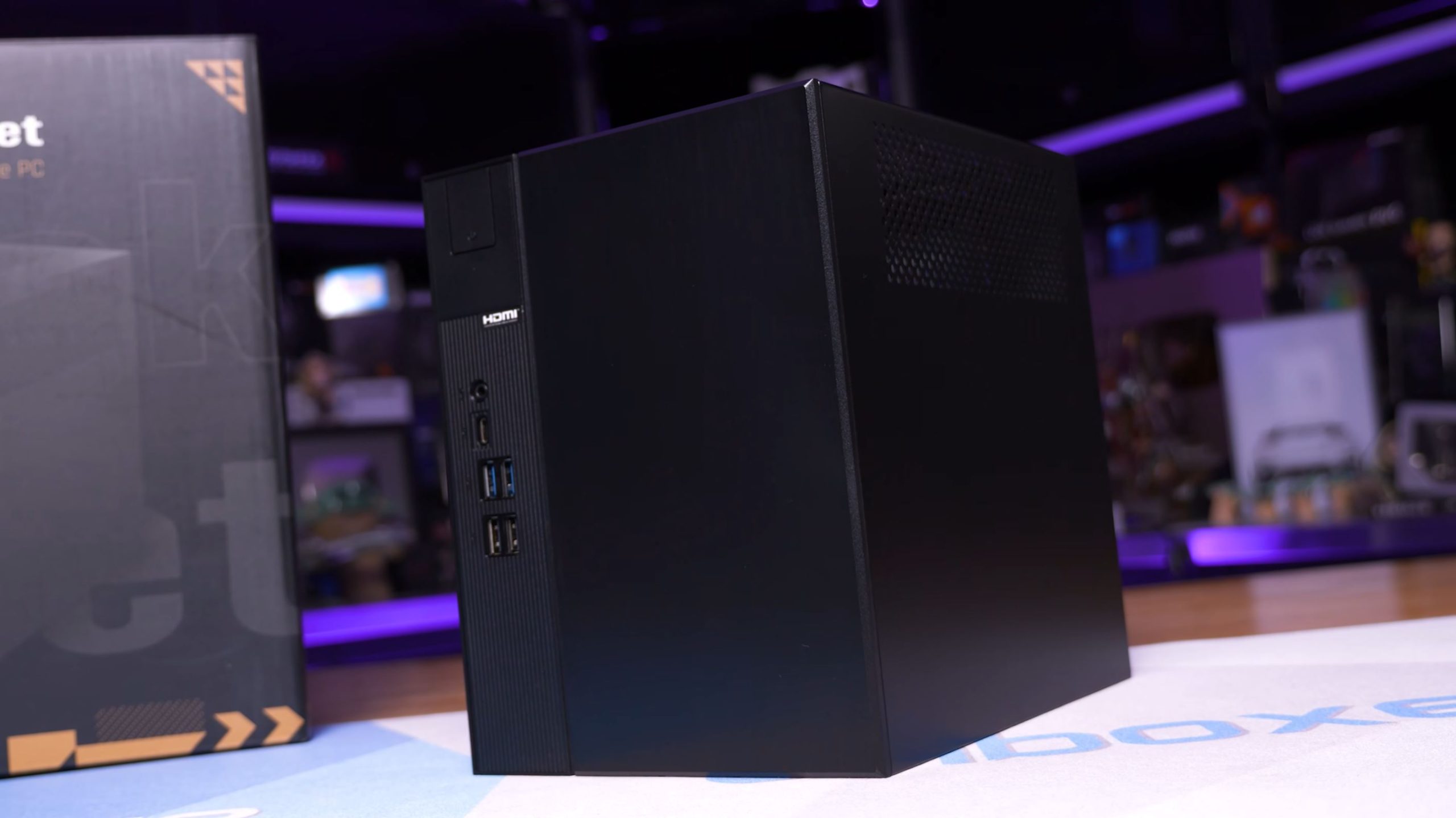 Un PC au format compact et noir posé à côté de son carton d'emballage, avec les ports HDMI, USB et audio visibles sur le panneau avant.
