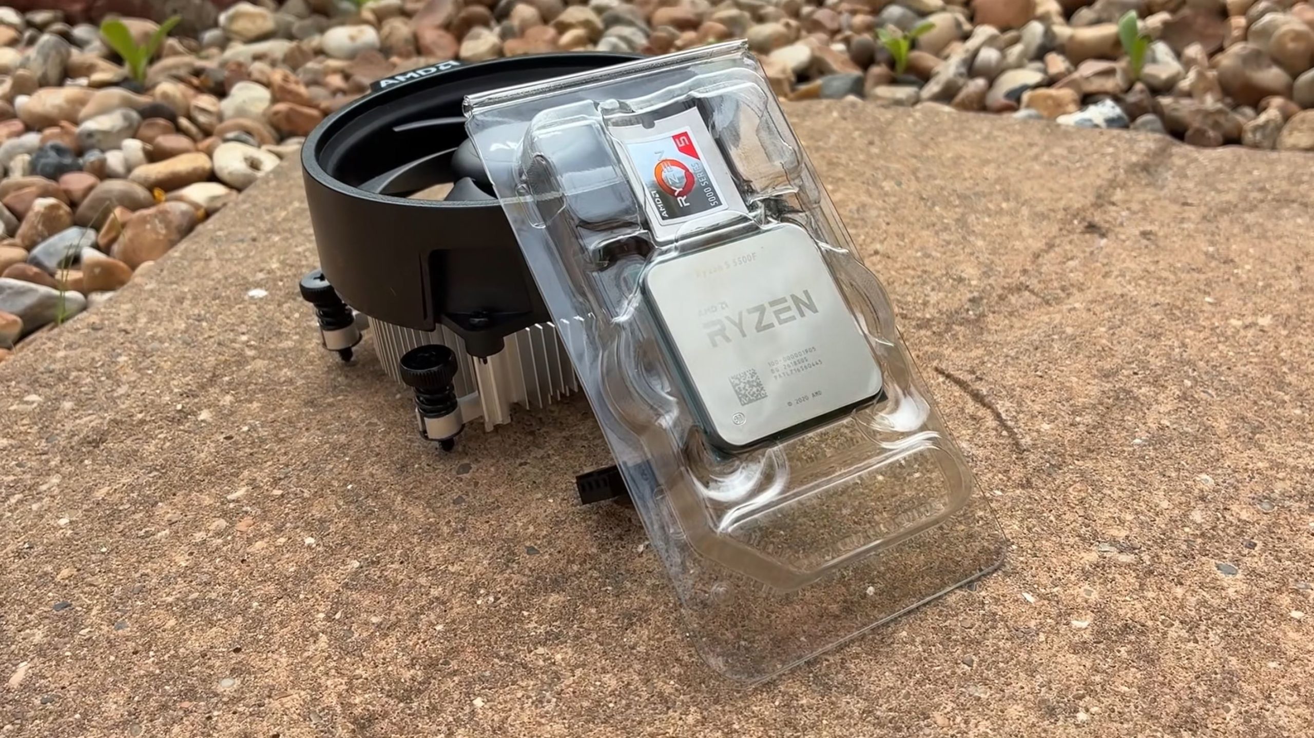Un processeur AMD Ryzen 5 5500F dans son emballage plastique posé sur une surface en pierre, avec un ventilateur AMD d'origine à côté.