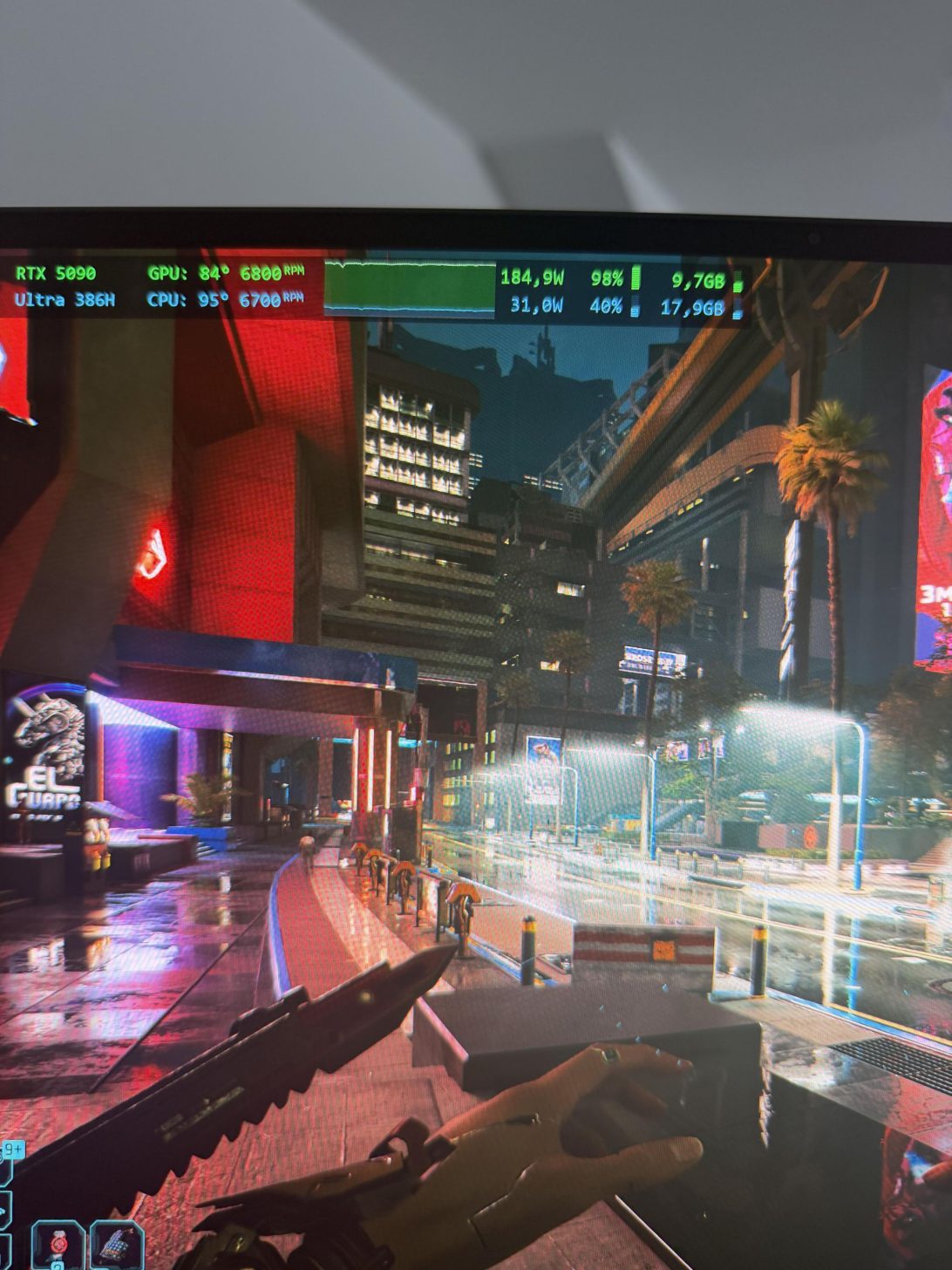 Une scène de gameplay de 'Cyberpunk 2077' montre un personnage tenant un couteau dans une rue néon. Les métriques à l'écran indiquent 'RTX 5090', une température GPU de '84°' et une température CPU de '95°'.