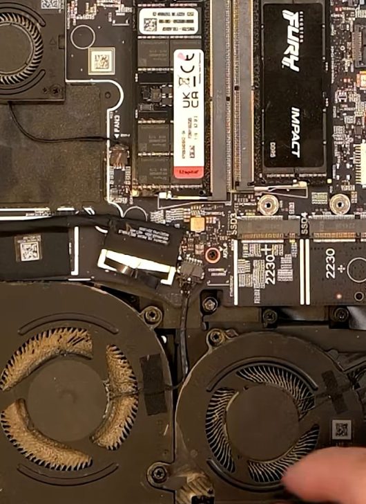 Vue interne d'un portable Alienware montrant la RAM et un SSD, avec des ventilateurs extrêmement encrassés.