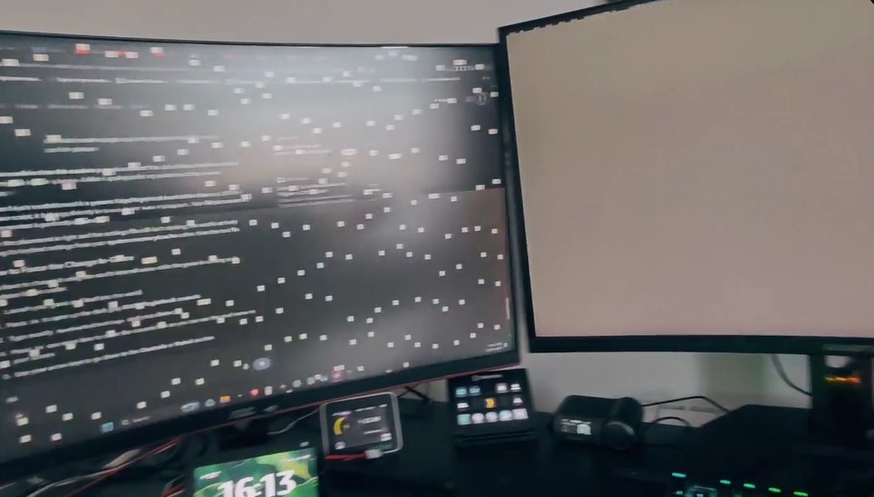 Deux écrans incurvés sur un bureau affichant un bureau en désordre et un écran vide, avec une horloge indiquant '16:13' sur un petit appareil en dessous.