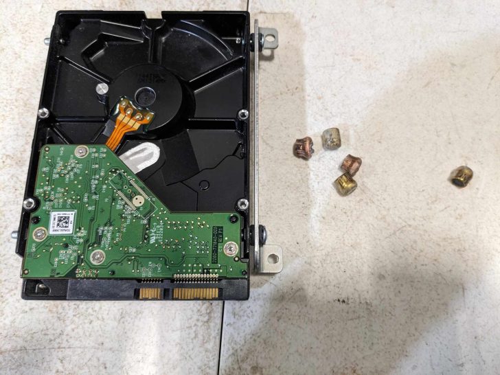 Un disque dur ouvert avec sa carte verte et ses plateaux visibles, posé à côté de cinq petits composants de couleur cuivre.