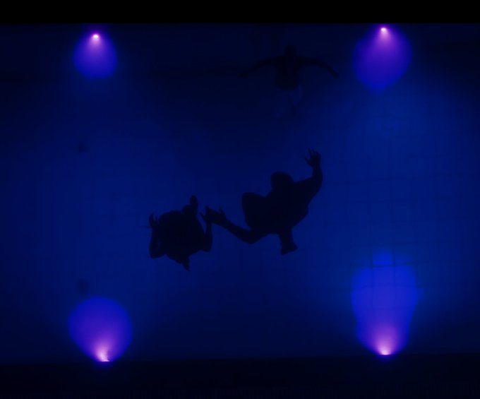 Trois silhouettes apparaissent sous l'eau, au sein d'une piscine éclairée par des lumières bleues.