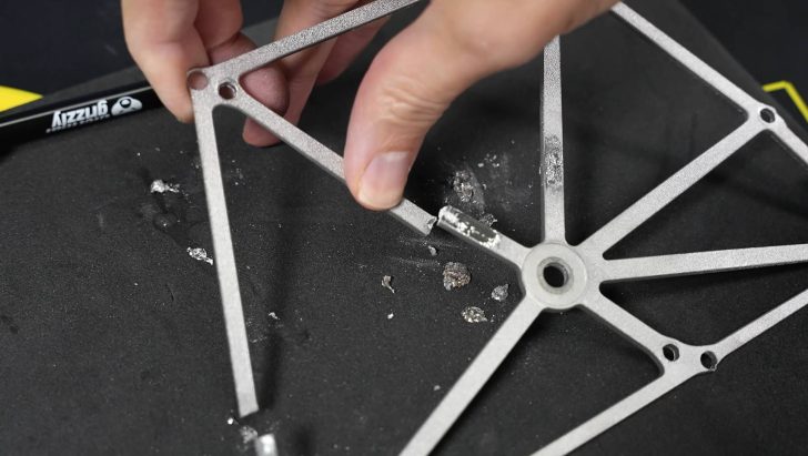 Une personne examine le cadre métallique cassé d'un composant, avec des débris visibles et un tournevis à proximité.