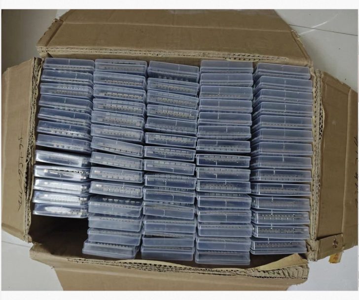 Une grande boîte en carton contenant des boîtiers plastiques transparents empilés et remplis de composants électroniques.