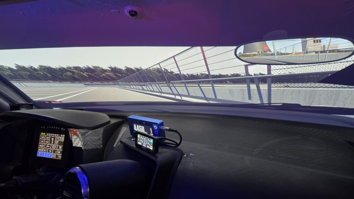 L'intérieur d'un simulateur de voiture de course montrant un compteur de vitesse numérique indiquant '4458' et une vue de circuit projetée sur le pare-brise.