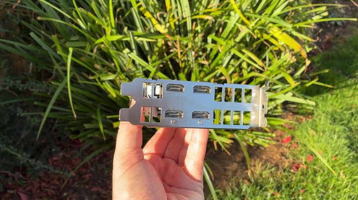 Une personne tient le connecteur d'une carte graphique avec trois ports DisplayPort et un port HDMI, sur un fond de verdure.