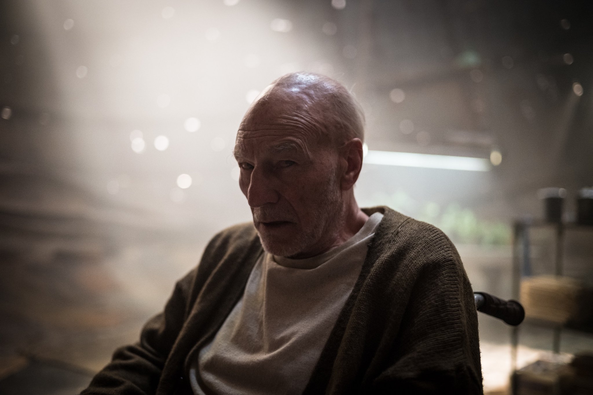 An older man in a dimly lit room, with a contemplative expression, sitting against a blurred background.