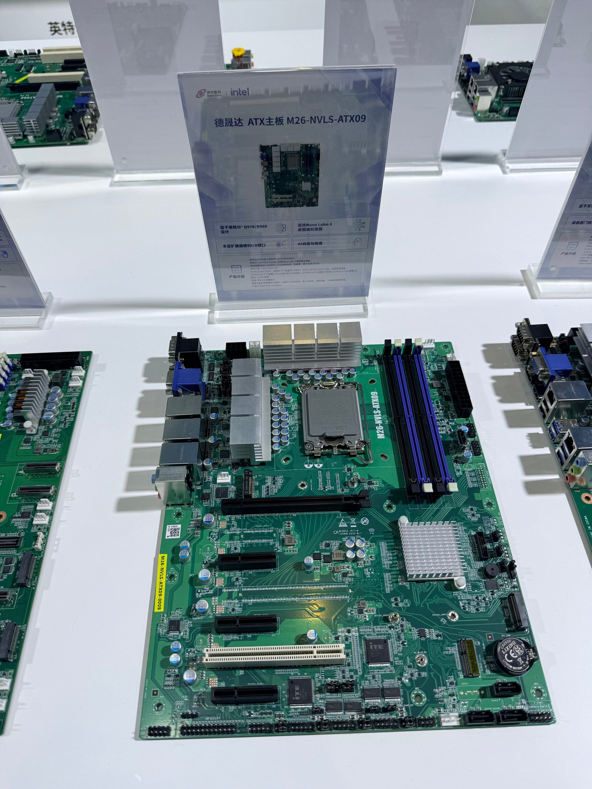 A Greenltd M26-NVLS-ATX09 motherboard with visible Intel branding is displayed on a table alongside a descriptive placard.