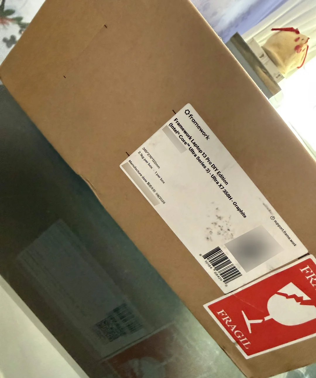 Un carton de livraison estampillé Fragile, contenant un Framework Laptop 13 DIY Edition Graphite avec processeur Intel Core Ultra Series 3.