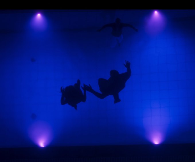 Deux silhouettes se dessinent sous l'eau, au sein d'une piscine baignée par des lumières bleues.